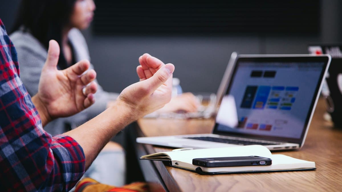 A person gesturing while talking across a table, with a laptop, notebook and phone in front of them