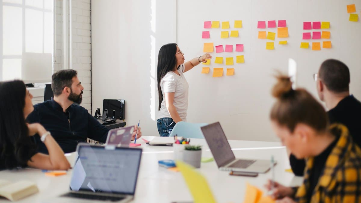 A person arranging sticky notes on a wall while colleagues with laptops watch from a meeting table