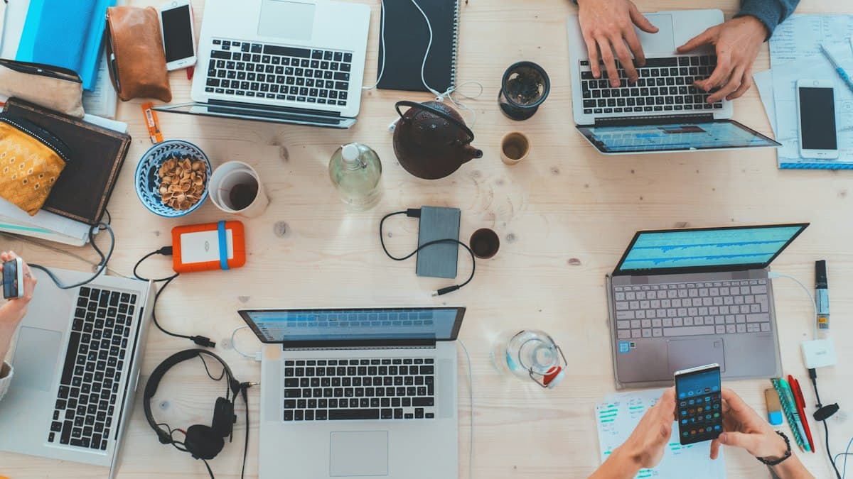 Top-down view of a shared wooden table with several laptops, phones and notebooks, with people working at it Top-down view of a shared wooden table with several laptops, phones and notebooks, with people working at it